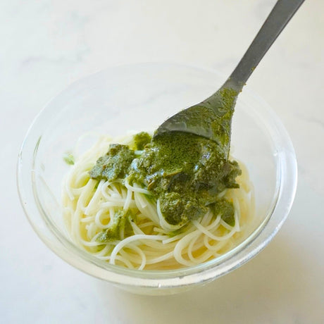 A clear glass bowl of somen noodles with a spoonful of green tea pesto sauce on a light marble surface.